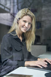 Portrait of smiling blond woman using laptop behind windowpane in office - PNEF02498