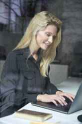 Smiling blond woman using laptop behind windowpane in office - PNEF02500