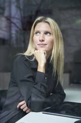 Portrait of blond woman with laptop behind windowpane in office - PNEF02501