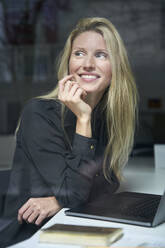 Portrait of happy blond woman with laptop behind windowpane in office - PNEF02502