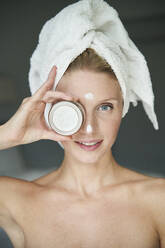 Porträt einer schönen Frau mit in ein Handtuch eingewickeltem Kopf, die sich ein Cremetöpfchen vor die Augen hält - PNEF02558