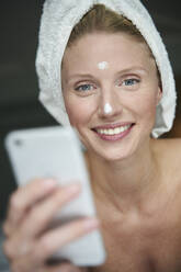 Porträt einer schönen Frau mit einem in ein Handtuch gewickelten Kopf, die ein Smartphone hält - PNEF02560