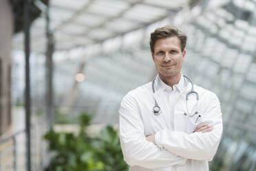 Portrait of doctor with stethoscope, standing in atrium, with arms crossed - JOSEF00150