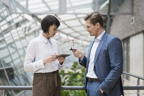 Geschäftsmann und Frau stehen im Atrium eines Bürogebäudes, benutzen ein digitales Tablet und halten einen Roboterarm - JOSEF00170