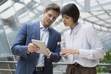 businessman and woman stnading in atrium of office building, using digital tablet, holding robot arm - JOSEF00171