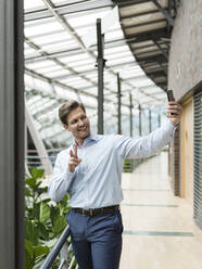 Businessman in green atrium, taking smartphone selfie, making victory sign - JOSEF00220