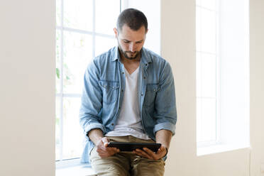 Young man at home looking at tablet sitting on window bench - SBOF02140