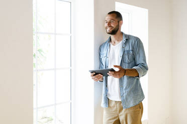 Young smiling man at home holding tablet standing at window and looking outside - SBOF02142