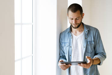 Young man at home smiling and looking at his tablet standing at window - SBOF02145