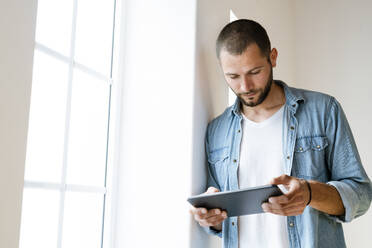 Young serious man at home looking at his tablet standing at window - SBOF02146