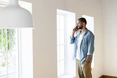 Junger Mann zu Hause im Wohnzimmer, der auf sein Smartphone spricht und aus dem Fenster schaut - SBOF02152