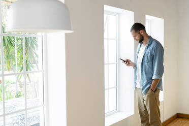 Junger Mann zu Hause, der sein Smartphone überprüft und vor seinen Fenstern im modernen Wohnzimmer steht - SBOF02153