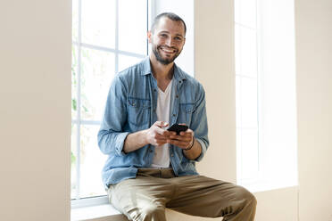 Happy young man at home holding his smartphone and sitting on window bench - SBOF02156