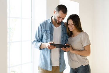 Lächelndes Paar zu Hause vor dem Fenster stehend, das gemeinsam ein Tablet betrachtet - SBOF02157