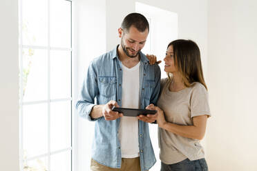 Smiling couple at home standing in front of window looking at tablet together - SBOF02158