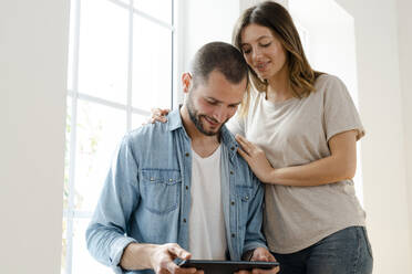 Smiling couple at home in front of window looking at tablet together - SBOF02159