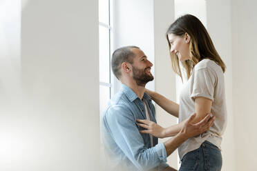 Affectionate smiling couple in love embracing each other at window in their home - SBOF02160