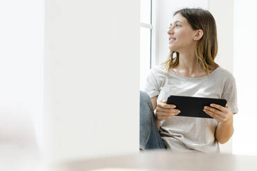 Young brunette woman at home sitting on window bench and looking outside while holding her tablet - SBOF02163