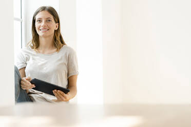 Smiling young brunette woman at home sitting on window bench and holding her tablet - SBOF02165