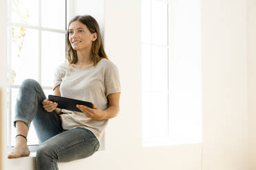 Lächelnde junge brünette Frau zu Hause sitzt auf Fensterbank und hält ihr Tablet - SBOF02166