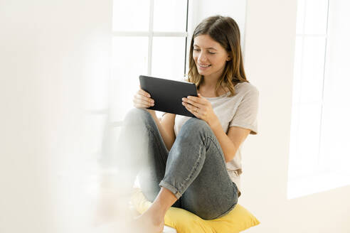 Lächelnde junge brünette Frau zu Hause sitzt auf Fensterbank und schaut auf ihr Tablet - SBOF02167