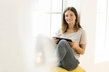 Smiling young brunette woman at home sitting on window bench and holding her tablet - SBOF02168