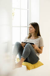 Serious young brunette woman at home sitting on window bench and holding her tablet while looking outside - SBOF02169