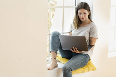 Junge brünette Frau zu Hause sitzt auf Fensterbank und hält ihren Laptop arbeiten im Home-Office - SBOF02171