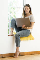 Lächelnde junge brünette Frau zu Hause sitzt auf Fensterbank und hält ihren Laptop arbeiten im Home-Office - SBOF02172