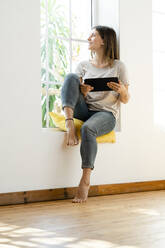 Smiling young brunette woman at home sitting on window bench and holding her tablet looking outside - SBOF02173