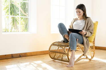 Smiling young brunette woman at home sitting in wooden armchair in front of window and looking at her tablet - SBOF02174