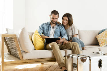 Lächelndes Paar zu Hause im modernen Wohnzimmer auf der Couch sitzend, während sie gemeinsam auf ein Tablet schauen - SBOF02189