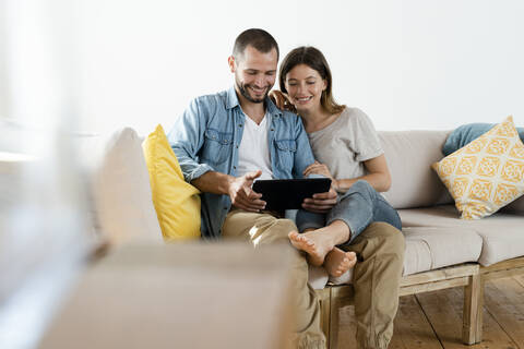 Glückliches Paar zu Hause im modernen Wohnzimmer auf der Couch sitzend, während sie gemeinsam auf ein Tablet schauen, lizenzfreies Stockfoto