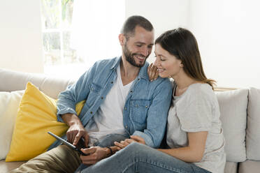 Glückliches Paar zu Hause im modernen Wohnzimmer auf der Couch sitzend, während sie gemeinsam auf ein Tablet schauen - SBOF02191