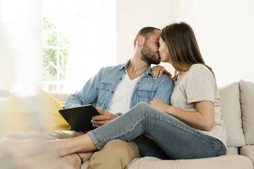 Affectionate couple relaxing at home and kissing while sitting on couch and holding tablet - SBOF02192