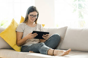 Smiling brunette woman with glasses relaxing on couch and looking at her tablet - SBOF02193