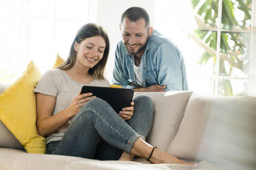 Glückliches Paar zu Hause im modernen Wohnzimmer auf der Couch und schaut gemeinsam auf ein Tablet - SBOF02195