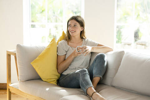 Lächelnde junge Frau, die sich zu Hause auf der Couch entspannt und einen Kaffeebecher hält, lizenzfreies Stockfoto