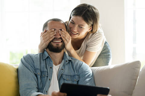 couple at home having fun together while she is covering his eyes and surprising him and he relaxes on couch holding tablet, lizenzfreies Stockfoto