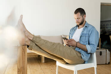 Junger Mann zu Hause tippt auf seinem Smartphone im modernen Wohnzimmer mit den Füßen auf dem Tisch - SBOF02214