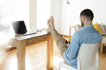 Young man at home typing on his smartphone in modern living room with his feet on the table - SBOF02215