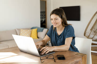 Lächelnde junge Frau, die von zu Hause aus im Home Office im modernen Wohnzimmer am Laptop arbeitet - SBOF02223