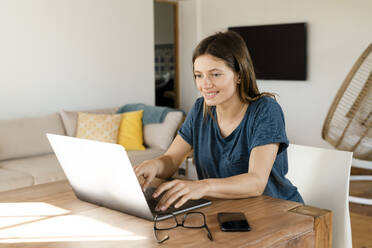 Smiling young woman working at laptop from home in home office in modern living room - SBOF02223