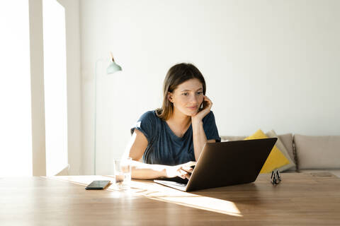 Zufriedene junge Frau arbeitet zu Hause am Laptop im modernen Wohnzimmer, lizenzfreies Stockfoto