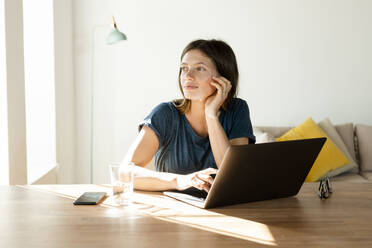 Junge Frau arbeitet zu Hause am Laptop im Büro in einem modernen Wohnzimmer und schaut nach draußen - SBOF02230