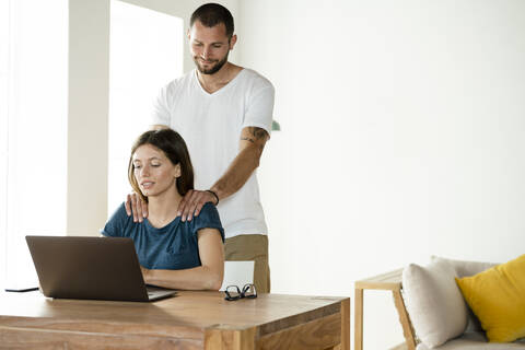 Frau arbeitet am Laptop, während ihr Freund ihr zu Hause den Nacken massiert, lizenzfreies Stockfoto