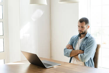 Serious young man with crossed arms at home looking at his laptop in home office - SBOF02251