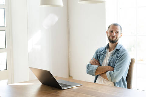 Lächelnder junger Mann mit verschränkten Armen bei der Arbeit an seinem Laptop im Büro zu Hause, lizenzfreies Stockfoto