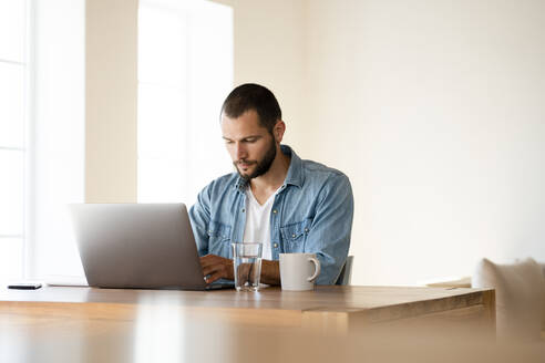 Konzentrierter junger Mann arbeitet zu Hause an seinem Laptop im Heimbüro im modernen Wohnzimmer - SBOF02262