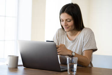 Lächelnde junge Frau zu Hause mit Laptop, die ihr Smartphone im Heimbüro überprüft - SBOF02268